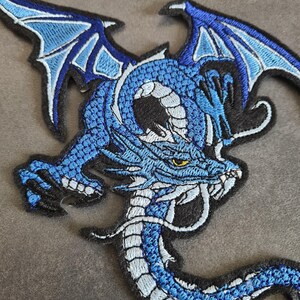 Könnte beinhalten: Gestickter Aufnäher mit einem blauen Drachen mit detaillierten Flügeln und Schuppen. Der Drache hat einen weißen Bauch und ein gelbes Auge, vor schwarzem Hintergrund. Der Aufnäher ist für das Aufnähen auf Kleidung oder Accessoires konzipiert.