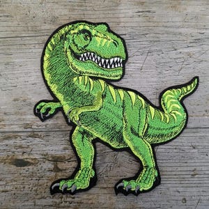 Puede incluir: Parche bordado con un dinosaurio Tyrannosaurus Rex verde con detalles amarillos. El dinosaurio tiene dientes blancos y garras negras, sobre un fondo de madera.