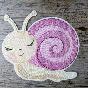 Könnte beinhalten: Gestickter Aufnäher in Form einer Schnecke mit cremefarbenem Kopf, geschlossenen Augen und einem lächelnden Mund. Das Gehäuse ist eine Spirale in Rosa- und Lilatönen. Zum Aufnähen oder Aufbügeln auf Stoff.