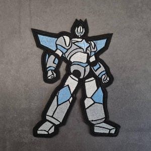Könnte beinhalten: Gestickter Aufnäher mit einem stilisierten Roboterdesign. Der Roboter ist hauptsächlich grau und weiß, mit blauen Akzenten an Schultern, Beinen und Brust. Er hat eine schwarze Umrandung und einen Stern auf der Brust.