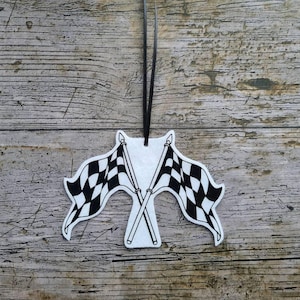 Könnte beinhalten: Ein weiß-schwarzes kariertes Flaggenornament mit zwei gekreuzten Flaggen. Das Ornament hängt an einem schwarzen Band vor einem rustikalen Holzgrund. Die Flaggen sind ein klassisches Rennsymbol.