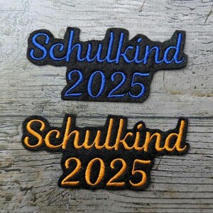 Könnte beinhalten: Zwei schwarze, gestickte Aufnäher mit dem deutschen Wort "Schulkind" und der Jahreszahl "2025". Einer hat blaue, der andere orangefarbene Buchstaben. Aufnäher auf Holzgrund.