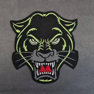 Könnte beinhalten: Schwarzer Panther-Aufnäher mit grimmigem Ausdruck. Der Kopf des Panthers ist neongrün umrandet, mit weißen Augen und roten Zähnen. Der Aufnäher befindet sich auf grauem Hintergrund.