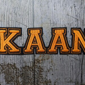 Op de afbeelding: Geborduurde patch met de naam "KAAN" in oranje blokletters met een zwarte rand. De patch ligt op een verweerde, grijze houten ondergrond. De letters hebben een klassieke, collegiale stijl.