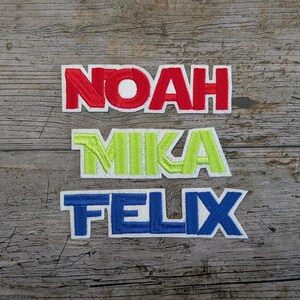 May include: Embroidered name patches: "NOAH" in red, "MIKA" in green and yellow, and "FELIX" in blue. Each name is in a bold, blocky font with a white border, set against a weathered wood background.