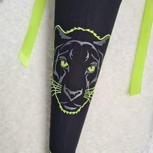 Könnte beinhalten: Schwarze, kegelförmige Tasche mit einem Panthergesicht-Design. Das Gesicht des Panthers ist in Grau und Neongrün gestickt. Neongrüne Bänder sind oben befestigt. Die Tasche ist wahrscheinlich für Schulmaterialien.