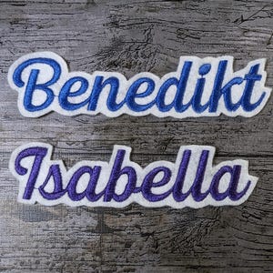 May include: Two embroidered name patches: "Benedikt" in blue script with a white border, and "Isabella" in purple script with a white border. The patches are on a weathered wood background.