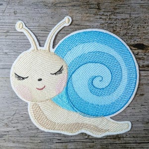 Könnte beinhalten: Gestickter Schneckenaufnäher mit hellblauem Gehäuse mit Spiralmuster. Die Schnecke hat einen cremefarbenen Körper, geschlossene Augen und einen lächelnden Mund. Der Aufnäher befindet sich auf einer Holzoberfläche.