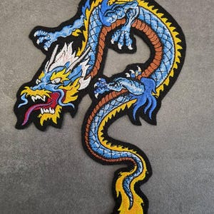 Könnte beinhalten: Gestickter Drachenaufnäher in Blau-, Gelb-, Orange- und Schwarztönen. Der Drache weist komplizierte Details auf, darunter eine rote Zunge und weiße Akzente. Der Aufnäher hat einen schwarzen Rand und ist wahrscheinlich für Kleidung oder Accessoires.