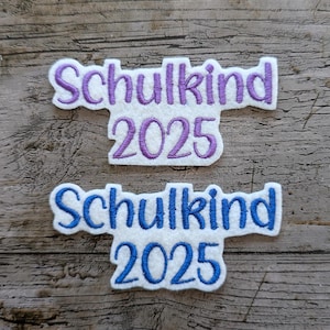 May include: Two white fabric patches with embroidered text. The top patch has purple text that reads "Schulkind 2025". The bottom patch has blue text that reads "Schulkind 2025". The patches are on a wooden surface.