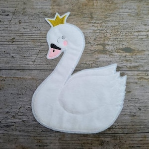 Może przedstawiać: Biały filcowy łabędź ze złotą koroną, różowymi policzkami i czarnym dziobem. Łabędź to fantazyjny design, idealny do dekoracji pokoju dziecięcego. Łabędź jest umieszczony na drewnianej powierzchni.