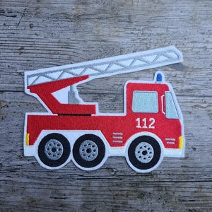 May include: Embroidered patch of a red fire engine with a white ladder, black wheels, and the number 112. The patch has a white border and is set against a wood grain background.