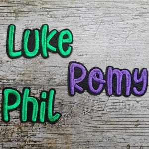 May include: Embroidered patches with the names "Luke", "Remy", and "Phil". The names are in a playful, rounded font, with green and purple stitching on a black background. The patches are on a weathered, wooden surface.