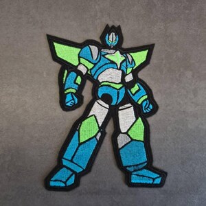 May include: Embroidered patch depicting a robot in blue, green, grey, and black. The robot has a geometric design with angular shapes and a star on its chest. The patch is designed for application on clothing or accessories.
