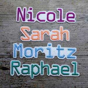 May include: Embroidered name patches in purple, orange, blue, and green. The names "Nicole", "Sarah", "Moritz", and "Raphael" are displayed on a weathered wood background. Each name is outlined in white.