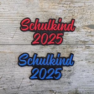 May include: Two embroidered patches with the text "Schulkind 2025". One patch has red lettering with a black border, and the other has blue lettering with a black border. The patches are on a weathered wooden surface.