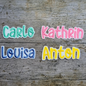 May include: Four embroidered name patches: "Carlo" in teal, "Kathrin" in pink, "Louisa" in blue, and "Anton" in yellow. Each name is outlined in white, set against a wooden background.