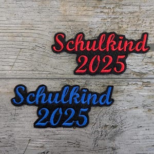 May include: Two embroidered patches with the text "Schulkind 2025". One patch has red lettering with a black border, and the other has blue lettering with a black border. The patches are on a weathered wooden surface.