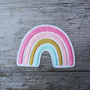 Könnte beinhalten: Gestickter Regenbogen-Aufnäher mit rosa, hellrosa, goldfarbenen und hellblauen Bögen. Der Aufnäher hat einen weißen Rand und ist vor einem grauen Holzgrund platziert. Ideal zum Aufnähen auf Kleidung oder Accessoires.