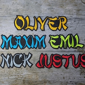 May include: Embroidered name patches: OLIVER in yellow and gold, MAXIM in blue, EMIL in green, NICK in white, and JUSTUS in red. The patches are on a dark grey background, with a wooden background.
