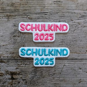 May include: Two embroidered patches with the German word "SCHULKIND" and the year "2025". One patch has pink text on a white background, the other has blue text on a white background. The patches are on a wooden surface.