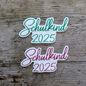 May include: Two white felt patches with the German word "Schulkind" and the year "2025" embroidered in green and pink. The patches are on a weathered wooden surface.
