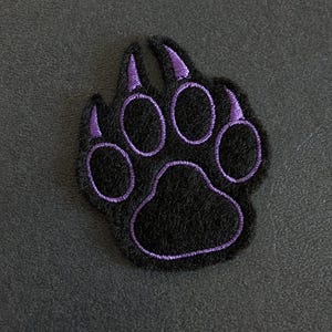 以下が含まれることがあります： 紫色の刺繍が施された黒い足跡パッチ。パッチは、個々のつま先パッドと爪を備えたクマの足の細部を描いています。デザインは暗い背景に配置され、印象的なコントラストを生み出しています。