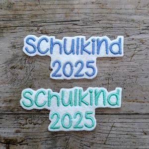 May include: Two embroidered patches with the text "Schulkind 2025" in blue and teal. The patches are white with a blue or teal outline and are set against a wooden background.