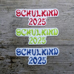 May include: Three embroidered patches with the text "SCHULKIND 2025" in red, green, and blue. The patches are white with a curved rectangular shape and are set against a weathered wood background.