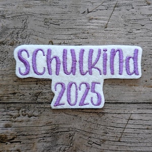 May include: A white felt patch with purple embroidered text that reads "Schulkind 2025". The patch has a rectangular shape with a small rectangular extension below the main text. The background is a weathered wood surface.