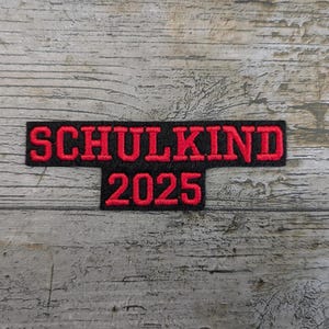 Könnte beinhalten: Ein schwarzer, gestickter Aufnäher mit dem roten Text "SCHULKIND 2025". Der Aufnäher ist rechteckig mit einem kleineren Rechteck darunter. Der Text ist in einer fetten, serifenlosen Schriftart. Der Hintergrund ist eine verwitterte, graue Holzoberfläche.