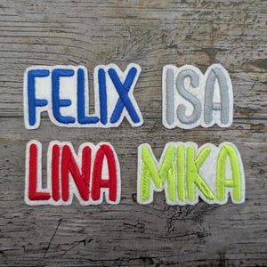 May include: Four embroidered name patches: "FELIX" in blue, "ISA" in gray, "LINA" in red, and "MIKA" in lime green. Each name is outlined in white, set against a wooden background.