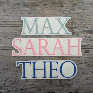May include: Three embroidered name patches: "MAX" in light blue, "SARAH" in pink, and "THEO" in blue. Each name is stitched on a white felt rectangle. The patches are displayed on a weathered, grey wooden surface.