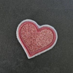 Könnte beinhalten: Ein herzförmiger Aufnäher mit einem glitzernden rosa Zentrum, umrandet von einem rosa gestickten Rand und einer weißen Filzkante. Der Aufnäher ist für dekorative Zwecke bestimmt, möglicherweise für Kleidung oder Accessoires.