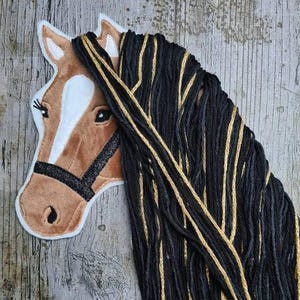 Op de afbeelding: Een paardenhoofd applicatie met een bruin en wit gezicht, zwarte teugel en lang zwart en goud garen haar. De applicatie is geplaatst tegen een verweerde houten achtergrond.