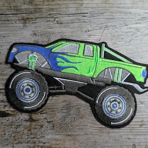 May include: Embroidered patch of a monster truck with large tires. The truck features a blue cab with green flames and a green bed. The tires and chassis are black, with gray and blue accents. The patch is designed for application on fabric.