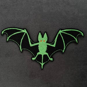 Può includere: Toppa ricamata a forma di pipistrello nero con contorni verdi e occhi arancioni. Le ali sono spiegate. Ideale per progetti a tema Halloween o gotico. Sfondo grigio scuro.