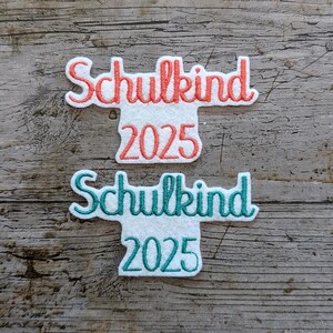 May include: Two white felt appliques with the German word "Schulkind" and the year "2025" in red and teal script. The appliques are on a weathered wood surface.