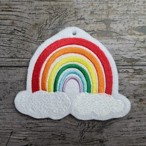 Könnte beinhalten: Ein farbenfrohes, regenbogenförmiges Applikationsmotiv mit gestickten Bögen in Rot, Orange, Gelb, Grün und Lila. Die Applikation ist auf weißem Filz, mit zwei weißen Wolkenformen unten. Für Bastelarbeiten.