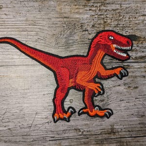 Könnte beinhalten: Gestickter Aufnäher mit einem roten und orangefarbenen Dinosaurier, wahrscheinlich einem Velociraptor, mit scharfen Zähnen und Krallen. Der Aufnäher hat einen schwarzen Rand und befindet sich auf einem Holzgrund. Geeignet für Kleidung oder Accessoires.