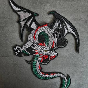 Könnte beinhalten: Gestickter Drachenaufnäher mit detailliertem Design. Der Drache hat einen grauen Körper, rote Akzente und grüne Schuppen. Seine Flügel sind schwarz und grau, und er hat gelbe Augen. Dieser Aufnäher eignet sich für Jacken, Taschen oder andere Stoffartikel.