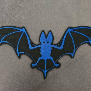May include: Embroidered bat patch featuring a black bat with blue wing details and a blue body. The bat has outstretched wings and is centered on a gray background. This patch is suitable for clothing or accessories.