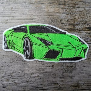 Könnte beinhalten: Gestickter Aufnäher eines grünen Sportwagens mit schwarzen Akzenten und weißem Rand. Das Auto-Design hat ein niedriges Profil und detaillierte Räder. Der Aufnäher liegt auf einer verwitterten Holzoberfläche.