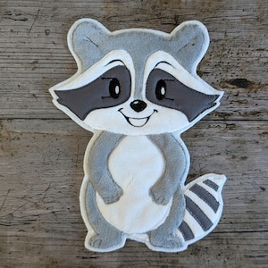 Könnte beinhalten: Ein Plüsch-Waschbär-Applikation in Grau-, Weiß- und Schwarztönen. Der Waschbär hat ein lächelndes Gesicht mit dunklen Augen und einen gestreiften Schwanz. Die Applikation ist zum Aufnähen auf Stoff gedacht.