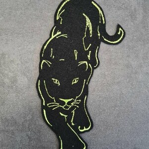 May include: Black panther patch with neon green embroidered detailing. The patch features a stylized panther design, with the animal's form outlined in bright green thread. The panther is depicted in a walking pose, with its head and body facing forward.