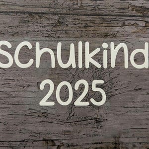 May include: White text on a wood-grain background reads "Schulkind 2025". The text is in a simple, rounded font. The background has a rustic, weathered appearance with various shades of gray and brown.