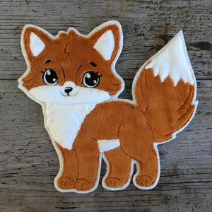 Könnte beinhalten: Ein orange-weißes Fuchsapplikation. Der Fuchs hat große, ausdrucksstarke Augen, eine weiße Brust und einen flauschigen Schwanz. Die Applikation ist wahrscheinlich für ein Bastelprojekt oder zum Dekorieren von Kleidung gedacht.