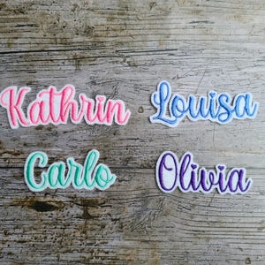 May include: Four embroidered name patches: "Kathrin" in pink, "Louisa" in blue, "Carlo" in teal, and "Olivia" in purple. Each name is in a cursive font with a white border, set against a wood grain background.