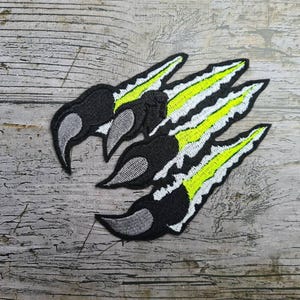 May include: Embroidered claw patch with a black claw design, grey accents, and a vibrant yellow and white stripe pattern. The patch is set against a rustic wooden background, suitable for jackets or bags.