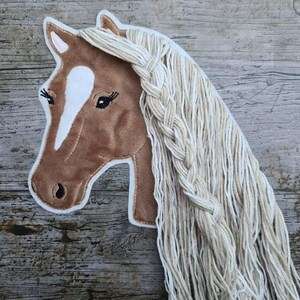 Op de afbeelding: Een bruine paardenhoofd wanddecoratie met een witte bles en een lange, gevlochten manen. De paardenkop is gemaakt van een zacht materiaal en is omrand met witte stiksels. De manen zijn gemaakt van lange, crèmekleurige strengen.